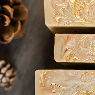 Close up of three bars of Fireside Mountain Retreat cold process soap on a wooden background surrounded by pine cones
